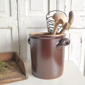 Puede incluir: Un recipiente de cerámica marrón para utensilios de cocina con dos asas, que contiene un batidor de alambre y cucharas de madera. El recipiente cilíndrico se encuentra sobre una superficie blanca. Una bandeja de madera con vegetación está en primer plano.