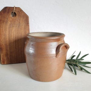 Peut inclure: Un pot en grès brun avec deux poignées et des lignes gravées horizontales. Une planche à découper en bois et une branche de feuilles vertes sont en arrière-plan. Un article de cuisine rustique et vintage.