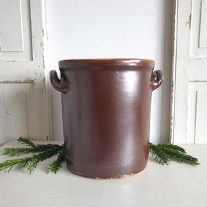 Könnte beinhalten: Ein brauner Keramiktopf mit zwei seitlichen Griffen. Der zylindrische Behälter hat einen leicht ausgestellten Rand und eine dunkelbraune Glasur. Der Topf steht auf einer weißen Oberfläche mit grünen Zweigen.