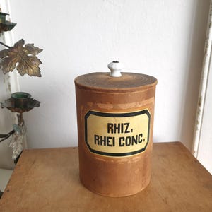 May include: A cylindrical brown cardboard apothecary jar with a removable lid featuring a small white ceramic knob. The front label is cream-colored with a black border and the black text reads RHIZ. RHEI CONC. The container has a weathered texture.