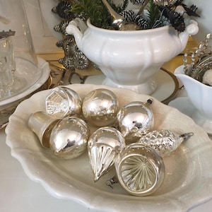 May include: A selection of antique silver Christmas baubles in diverse forms, such as bells, spheres, and teardrops, presented on a white, scalloped-edge dish. Other white ceramic serving dishes and decorative items are in the background.
