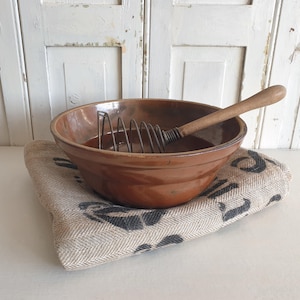 Könnte beinhalten: Eine braune Keramik-Rührschüssel mit einem Schneebesen und Holzgriff liegt auf einem beige-schwarz gemusterten Tuch. Die Schüssel hat eine glänzende Oberfläche, ideal zum Backen oder Kochen.