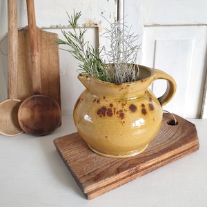 Può includere: Una brocca in ceramica giallo-marrone con manico e beccuccio, decorata con macchie marrone scuro. Contiene rametti di piante verdi e grigio argento. La brocca poggia su un piccolo tagliere quadrato in legno. Altri utensili da cucina in legno sono sullo sfondo.