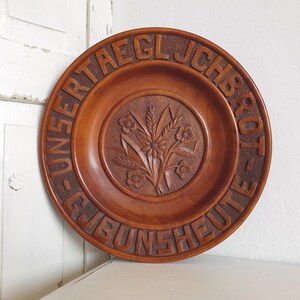 May include: A round, wooden plate with carved floral details in the center and embossed text around the rim. The plate is a warm brown color and appears to be vintage or antique.