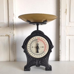 May include: A vintage black kitchen scale with a cream-colored face and a gold-toned bowl. The scale features a circular dial with black and red markings, and the word "FS" is visible. The scale is set against a white background.