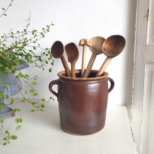 Puede incluir: Una vasija de cerámica esmaltada en color marrón oscuro con dos asas laterales que contiene un conjunto de cinco utensilios de cocina de madera, incluyendo varias cucharas y una herramienta de cabeza plana, colocados verticalmente.