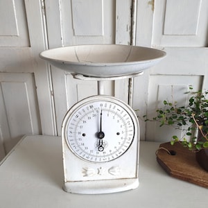 May include: A vintage white kitchen scale with a round face and a large, shallow bowl. The scale has a circular dial with black numbers and markings. The bowl is a light gray color. The scale is likely used for measuring ingredients.