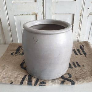 Puede incluir: Una olla de cerámica gris claro con una abertura ancha y un cuerpo ligeramente redondeado. La olla tiene una pequeña marca grabada en el lateral. Se encuentra sobre un saco de arpillera con texto negro. El fondo es una puerta de madera blanca desgastada.