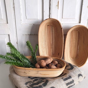 Op de afbeelding: Drie lichtbruine, ovaalvormige rotan manden van verschillende formaten. Eén mand bevat walnoten en groene takjes. De manden liggen op een patroonstof. De achtergrond is een witte wand met panelen.