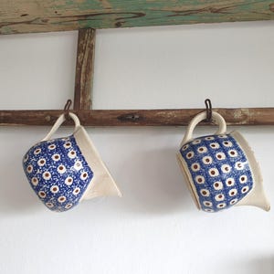 Op de afbeelding: Twee keramische mokken met blauw-witte bloempatronen hangen aan een rustieke houten balk. De mokken hebben een unieke vorm met een wit interieur en handvat. De mokken zijn opgehangen met metalen haken tegen een witte muur.