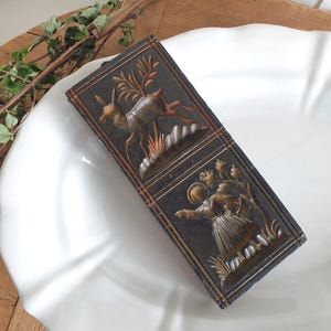 May include: A rectangular, dark brown, antique wooden box with two carved panels. One panel depicts a leaping deer, the other a person gathering flowers. The box is on a white plate.