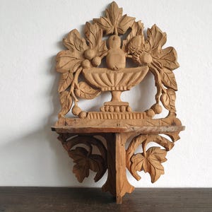 Könnte beinhalten: Ein hölzernes Wandregal mit einem geschnitzten Design mit Blättern, Früchten und einer zentralen Vogelfigur. Das Regal hat eine kleine, rechteckige Oberfläche zur Präsentation von Gegenständen und ist aus hellbraunem Holz gefertigt.