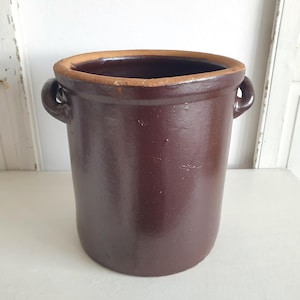 Può includere: Un vaso cilindrico in gres marrone scuro con bordo marrone chiaro e due piccole maniglie. Il contenitore antico è probabilmente utilizzato per la conservazione degli alimenti o la decorazione. Il vaso è su una superficie bianca.