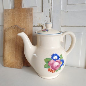 Op de afbeelding: Crèmekleurige keramische theepot met een bloemenontwerp met roze rozen, blauwe madeliefjes en rode bessen. De theepot heeft een deksel met een bijpassend bloemenpatroon en een blauwe rand. Een houten snijplank staat op de achtergrond.