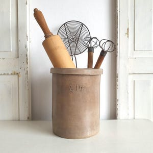 Può includere: Una collezione di utensili da cucina in un contenitore di ceramica marrone. Il contenitore ha il testo "4 Ltr" sul lato. Sono inclusi un mattarello di legno, una frusta e un colino a filo. Lo sfondo presenta porte bianche e una superficie bianca.