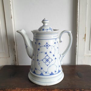 Peut inclure: Cafetière en porcelaine blanche avec un motif floral bleu. La cafetière a un couvercle, une anse incurvée et un bec verseur. Le motif comprend des motifs floraux et géométriques. La cafetière est posée sur une surface en bois foncé.