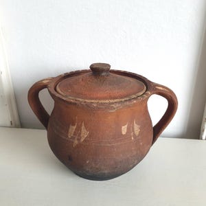 Könnte beinhalten: Ein brauner Keramiktopf mit Deckel und zwei Griffen. Der Topf hat eine runde Form und ist mit verblassten weißen Markierungen verziert. Der Deckel hat einen kleinen Knopf. Der Topf wird wahrscheinlich zum Kochen oder Aufbewahren verwendet.