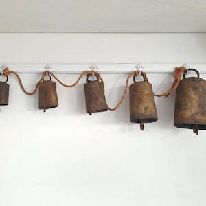 May include: A collection of five rustic metal bells of varying sizes, suspended from a white surface by a brown rope. The bells have a weathered, aged appearance with a tarnished, golden-brown hue. The rope is knotted between each bell.