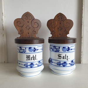 May include: A pair of ceramic spice jars with wooden lids. The jars are white with blue floral patterns and the words "Mehl" and "Salz" in black script. The wooden lids have a decorative carved design.