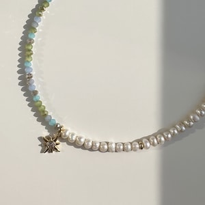 May include: A delicate necklace with a gold star charm. The necklace is made of small, light blue, green, and white beads. The star charm has a small clear stone in the center.