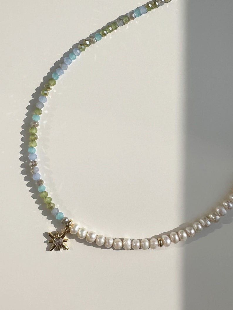 May include: A delicate necklace with a gold star charm. The necklace is made of small, light blue, green, and white beads. The star charm has a small clear stone in the center.