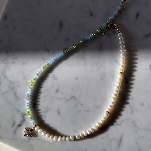 May include: A delicate necklace with a gold star charm, featuring a gradient of light blue, green, and white beads. The necklace is made with freshwater pearls and gemstone beads.