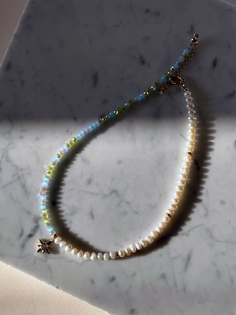 May include: A delicate necklace with a gold star charm, featuring a gradient of light blue, green, and white beads. The necklace is made with freshwater pearls and gemstone beads.