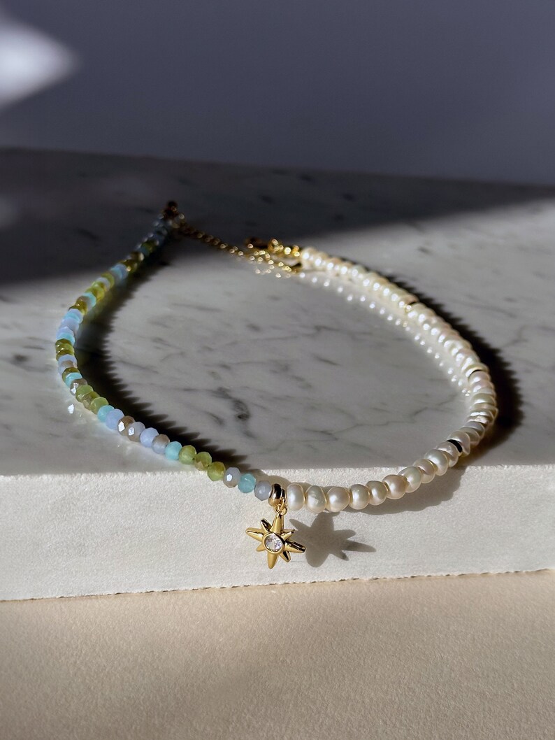 May include: A delicate gold necklace with a star charm. The necklace is made of small white pearls and green, blue, and clear glass beads.