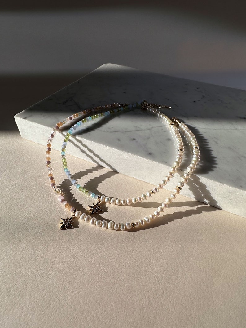 May include: Two delicate necklaces with starburst charms. One necklace is made of white pearls and the other is made of multi-colored beads.