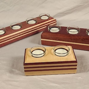 May include: Three wooden candle holders with multiple compartments for tealight candles. The holders are made of different types of wood, including a reddish-brown wood and a light-colored wood. The holders are arranged in a row on a white surface.