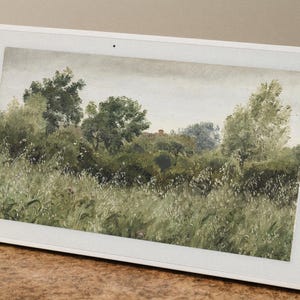 May include: A white framed digital display showing a landscape painting of a field and trees. The artwork features a muted color palette of greens, whites, and browns, with a cloudy sky. The display is angled on a brown surface.