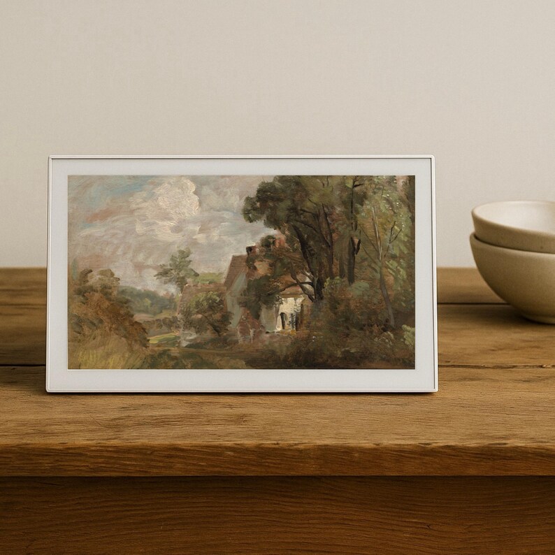 May include: A framed landscape painting depicting a rural scene with a house, trees, and a cloudy sky. The artwork is in a white frame and rests on a wooden surface. The painting's color palette includes greens, browns, and blues.