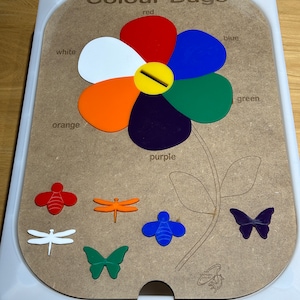 Könnte beinhalten: Ein Holzspielzeug mit einem farbzuordnenden Brett in Form einer Blume. Die Blume hat Blütenblätter in Rot, Weiß, Orange, Lila, Blau und Grün. Enthält farbige insektenförmige Teile. Der Text "Colour Bugs" befindet sich oben.