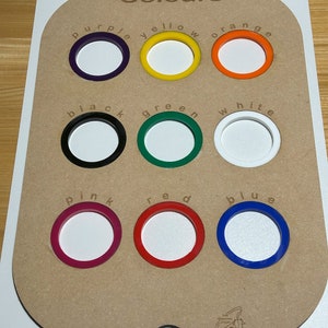 Könnte beinhalten: Ein Holzbrett mit neun kreisförmigen Ausschnitten, die jeweils mit einem Farbnamen und einem entsprechenden Farbring beschriftet sind. Die Farben sind lila, gelb, orange, schwarz, grün, weiß, rosa, rot und blau.