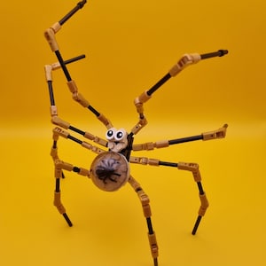 Puede incluir: Una araña Lego negra y dorada con grandes ojos blancos y un cuerpo transparente que muestra una araña negra en su interior. La araña tiene ocho patas y está posada sobre un fondo amarillo.