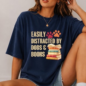 Peut inclure: Un t-shirt bleu marine à manches courtes avec le texte "EASILY DISTRACTED BY DOGS & BOOKS" en jaune clair. Le motif comprend deux empreintes de pattes en rouge et orange, ainsi qu'une pile de cinq livres colorés avec un livre ouvert au sommet.