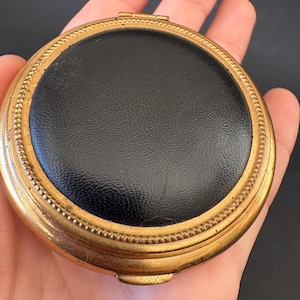 May include: A vintage compact mirror with a black leather center and a gold-toned metal frame. The frame has a decorative beaded edge. The compact is round and closed.