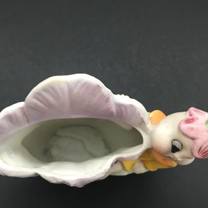 May include: A ceramic planter shaped like a girl with a pink flower on her head. The planter is painted with yellow, pink, and green details. The planter has a white interior and is designed to hold small plants.