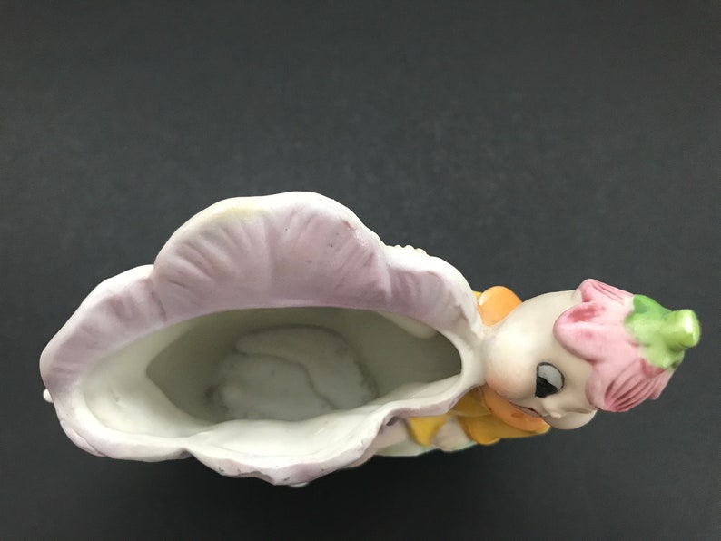 May include: A ceramic planter shaped like a girl with a pink flower on her head. The planter is painted with yellow, pink, and green details. The planter has a white interior and is designed to hold small plants.