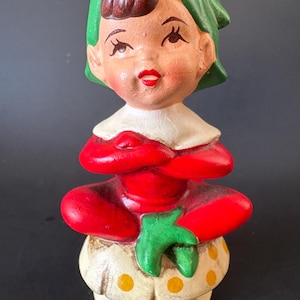 May include: A ceramic figurine of a seated elf with crossed arms. The elf wears a red dress, a green hat, and a white collar. The elf is sitting on a mushroom-shaped base with yellow polka dots. The figurine has rosy cheeks and red lips.