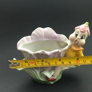 May include: A porcelain planter shaped like a flower with a whimsical fairy figure holding onto the side. The planter is painted in shades of pink, green, and yellow.