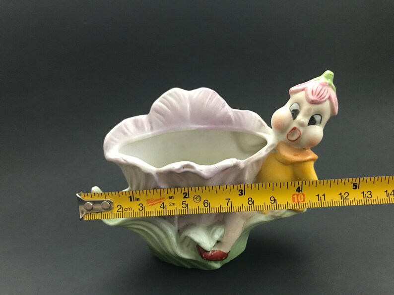 May include: A porcelain planter shaped like a flower with a whimsical fairy figure holding onto the side. The planter is painted in shades of pink, green, and yellow.