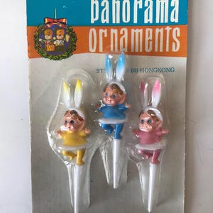 May include: Three pastel-colored bunny cake toppers with white bunny ears. The bunny toppers are wearing yellow, blue, and pink dresses. The package says "panorama ornaments" and "161 HONGKONG".