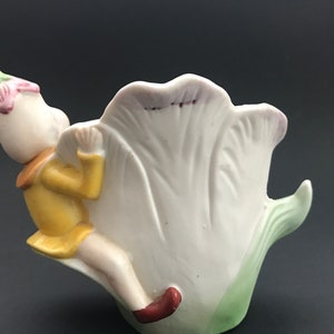 May include: A porcelain figurine of a fairy sitting on a white tulip. The fairy is wearing a yellow dress and a pink flower on its head. The tulip is green at the base and has a pink rim.