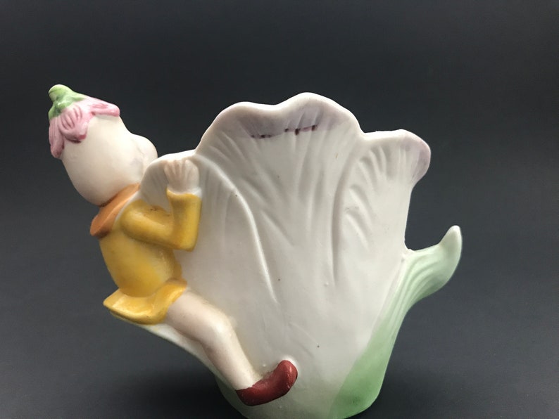 May include: A porcelain figurine of a fairy sitting on a white tulip. The fairy is wearing a yellow dress and a pink flower on its head. The tulip is green at the base and has a pink rim.