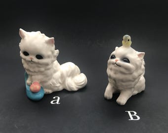 Puede incluir: Dos figuritas de gato de cerámica blanca. El gato de la izquierda sostiene una pelota azul y rosa. El gato de la derecha tiene un pequeño pájaro amarillo en la cabeza.