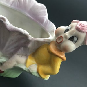 May include: A ceramic planter shaped like a fairy or gnome with a pink flower on its head. The planter is holding a flower pot and has a yellow shirt and red shoes.