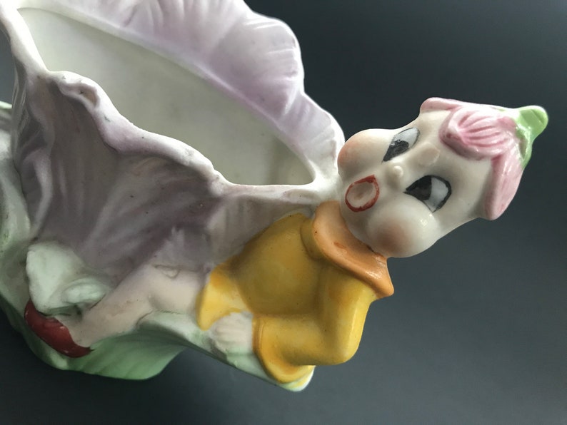 May include: A ceramic planter shaped like a fairy or gnome with a pink flower on its head. The planter is holding a flower pot and has a yellow shirt and red shoes.