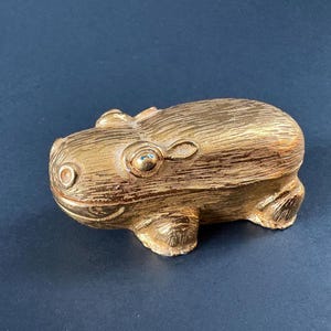 May include: A gold-colored, decorative frog figurine. The frog has a textured surface and is resting on a dark blue surface. The frog has large eyes and a smiling mouth. The figurine is a decorative item.