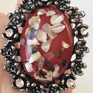 May include: A silver ring with a red and white oval stone set in a floral design. The stone is made of crushed shells and has a sparkly finish.